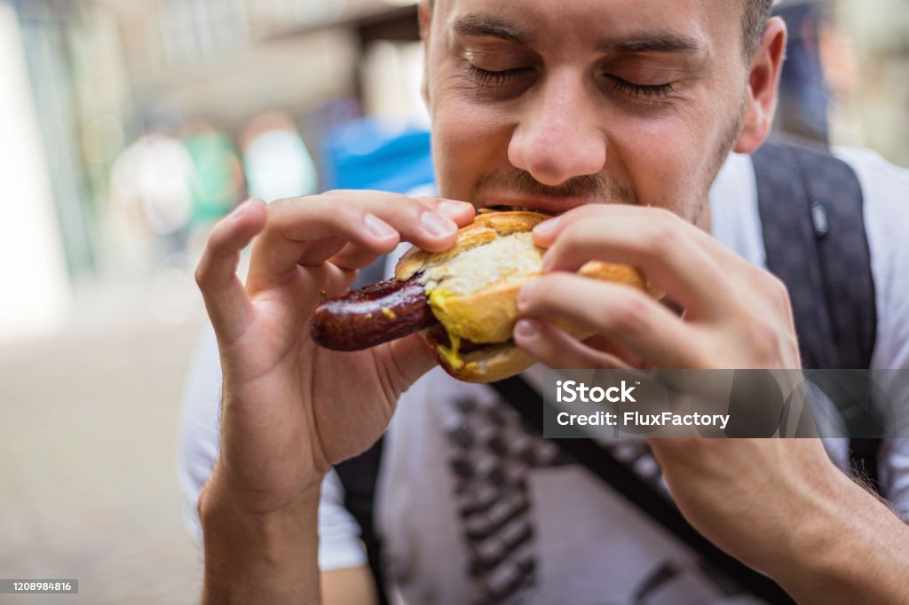 Hungry young male tourist eating German street food, bratwurst at the city street